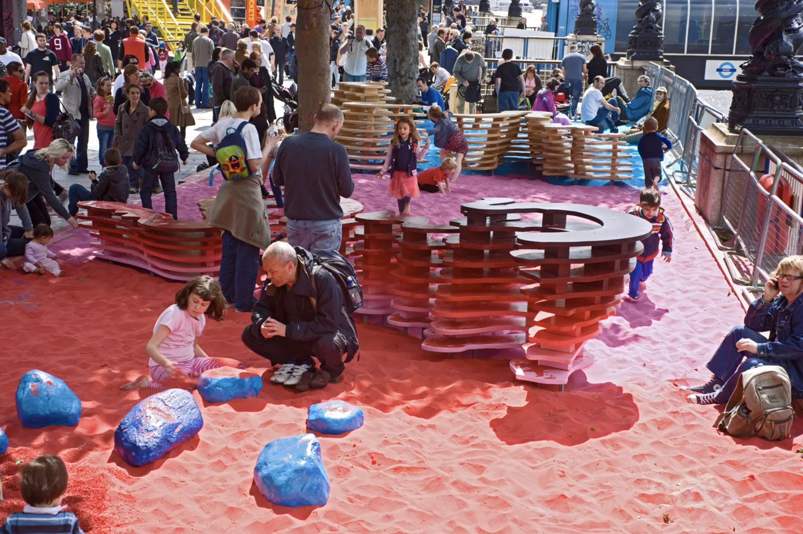 Visitors playing among coloured sand, tiered wooden structures and painted boulders at Rainbow Park on the South Bank, London.